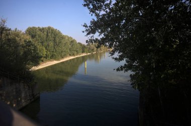 Sahili ve gökyüzünü ağaçların çerçevelediği ve yukarıdaki bir yol köprüsünden görülebilen suya dökmüş, saptırıcı bir kanalda demirleme direkleri.