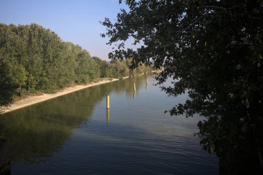 Sahili ve gökyüzünü ağaçların çerçevelediği ve yukarıdaki bir yol köprüsünden görülebilen suya dökmüş, saptırıcı bir kanalda demirleme direkleri.