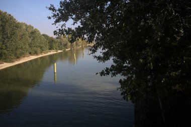 Sahili ve gökyüzünü ağaçların çerçevelediği ve yukarıdaki bir yol köprüsünden görülebilen suya dökmüş, saptırıcı bir kanalda demirleme direkleri.