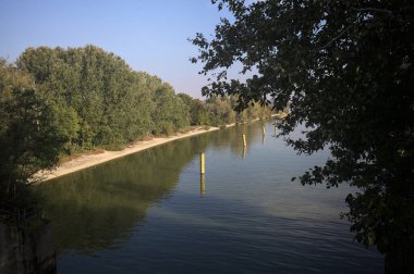 Sahili ve gökyüzünü ağaçların çerçevelediği ve yukarıdaki bir yol köprüsünden görülebilen suya dökmüş, saptırıcı bir kanalda demirleme direkleri.