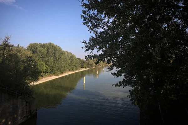 Sahili ve gökyüzünü ağaçların çerçevelediği ve yukarıdaki bir yol köprüsünden görülebilen suya dökmüş, saptırıcı bir kanalda demirleme direkleri.