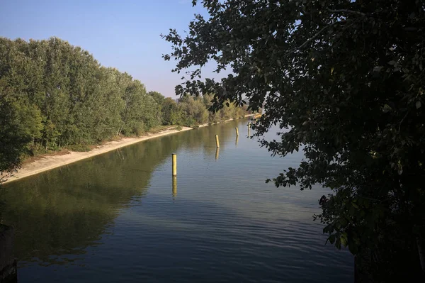Sahili ve gökyüzünü ağaçların çerçevelediği ve yukarıdaki bir yol köprüsünden görülebilen suya dökmüş, saptırıcı bir kanalda demirleme direkleri.