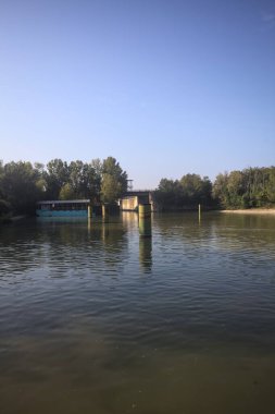 Kıyıların yanında demirleme direkleri ve güneşli bir günde İtalyan kırsalında ağaçlarla çevrili bir yol köprüsü olan saptırıcı bir kanal.