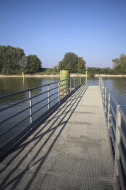 Oyalama kanalı üzerinde metalik tahta kaldırımlı iskele demirleme direklerinin yanında ve karşı kıyıda ağaçlarla çevrili.