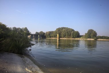 Kıyıların yanında demirleme direkleri ve güneşli bir günde İtalyan kırsalında ağaçlarla çevrili bir yol köprüsü olan saptırıcı bir kanal.