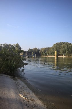 Kıyıların yanında demirleme direkleri ve güneşli bir günde İtalyan kırsalında ağaçlarla çevrili bir yol köprüsü olan saptırıcı bir kanal.
