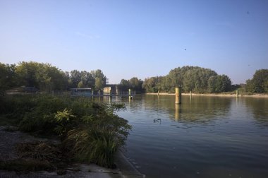 Kıyıların yanında demirleme direkleri ve güneşli bir günde İtalyan kırsalında ağaçlarla çevrili bir yol köprüsü olan saptırıcı bir kanal.