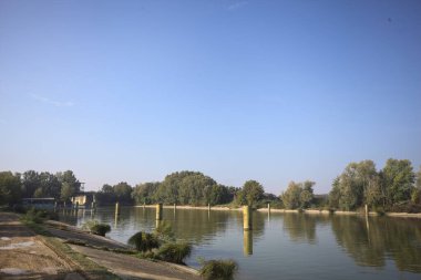 Kıyıların yanında demirleme direkleri ve güneşli bir günde İtalyan kırsalında ağaçlarla çevrili bir yol köprüsü olan saptırıcı bir kanal.