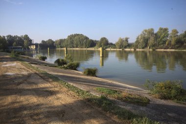 Kıyıların yanında demirleme direkleri ve güneşli bir günde İtalyan kırsalında ağaçlarla çevrili bir yol köprüsü olan saptırıcı bir kanal.