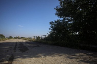 İtalya kırsalında güneşli bir günde, bir ucunda metalik korkuluklar olan yıpranmış bir köprü.