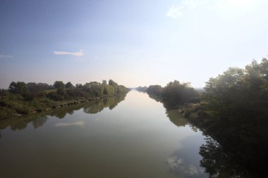 Nehir, üzerinde bir köprüden görünen suya yansıyan ağaçlarla çevrili güneşle kesişir.
