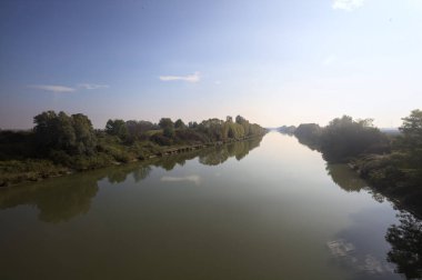 Nehir, üzerinde bir köprüden görünen suya yansıyan ağaçlarla çevrili güneşle kesişir.