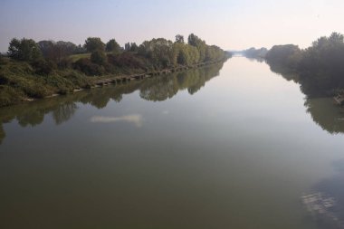 Nehir, üzerinde bir köprüden görünen suya yansıyan ağaçlarla çevrili güneşle kesişir.
