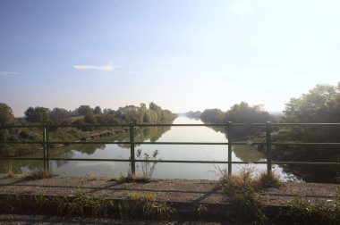 Nehir, üzerinde bir köprüden görünen suya yansıyan ağaçlarla çevrili güneşle kesişir.