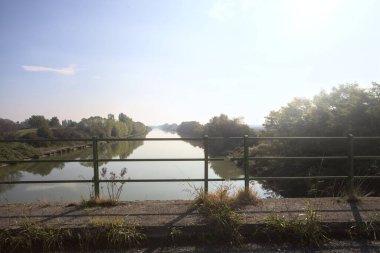 Nehir, üzerinde bir köprüden görünen suya yansıyan ağaçlarla çevrili güneşle kesişir.