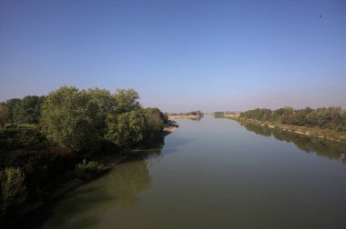 Ağaçlarla çevrili bir nehir, İtalya kırsalında güneşli bir günde su yüzeyine yansıyan bir setten görünüyor.