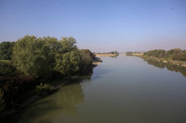 Ağaçlarla çevrili bir nehir, İtalya kırsalında güneşli bir günde su yüzeyine yansıyan bir setten görünüyor.