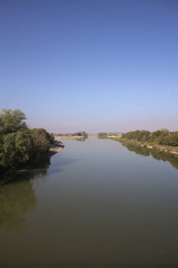Ağaçlarla çevrili bir nehir, İtalya kırsalında güneşli bir günde su yüzeyine yansıyan bir setten görünüyor.