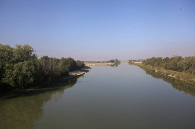 Ağaçlarla çevrili bir nehir, İtalya kırsalında güneşli bir günde su yüzeyine yansıyan bir setten görünüyor.