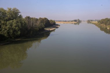 Ağaçlarla çevrili bir nehir, İtalya kırsalında güneşli bir günde su yüzeyine yansıyan bir setten görünüyor.