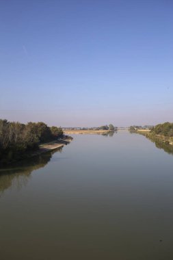 Ağaçlarla çevrili bir nehir, İtalya kırsalında güneşli bir günde su yüzeyine yansıyan bir setten görünüyor.