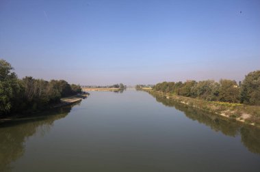 Ağaçlarla çevrili bir nehir, İtalya kırsalında güneşli bir günde su yüzeyine yansıyan bir setten görünüyor.