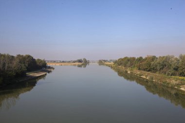 Ağaçlarla çevrili bir nehir, İtalya kırsalında güneşli bir günde su yüzeyine yansıyan bir setten görünüyor.