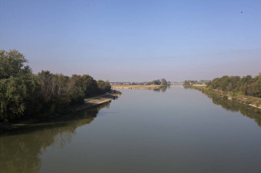 Ağaçlarla çevrili bir nehir, İtalya kırsalında güneşli bir günde su yüzeyine yansıyan bir setten görünüyor.