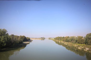 Ağaçlarla çevrili bir nehir, İtalya kırsalında güneşli bir günde su yüzeyine yansıyan bir setten görünüyor.
