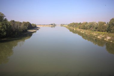 Ağaçlarla çevrili bir nehir, İtalya kırsalında güneşli bir günde su yüzeyine yansıyan bir setten görünüyor.