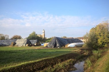 İtalya kırsalındaki bir köyün evleri güneşli bir günde bir tarlada balya yığınlarıyla çevrili bir kilisenin çan kulesi.