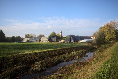İtalya kırsalındaki bir köyün evleri güneşli bir günde bir tarlada balya yığınlarıyla çevrili bir kilisenin çan kulesi.