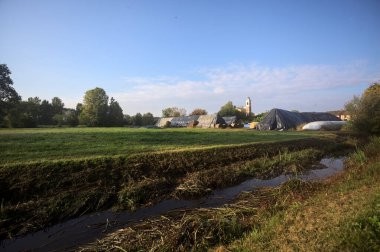 İtalya kırsalındaki bir köyün evleri güneşli bir günde bir tarlada balya yığınlarıyla çevrili bir kilisenin çan kulesi.