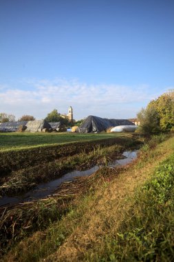 İtalya kırsalındaki bir köyün evleri güneşli bir günde bir tarlada balya yığınlarıyla çevrili bir kilisenin çan kulesi.
