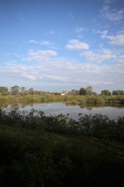Etrafı sazlıklarla çevrili bir gölet. Arkasında tarlalar ve uzakta evler var. Gökyüzü de suyun üzerine düşmüş.