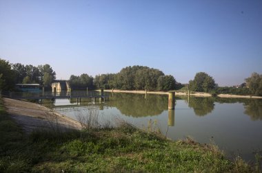 Kıyıların yanında demirleme direkleri ve güneşli bir günde İtalyan kırsalında ağaçlarla çevrili bir yol köprüsü olan saptırıcı bir kanal.