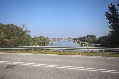 Sahili ve gökyüzünü ağaçların çerçevelediği ve yukarıdaki bir yol köprüsünden görülebilen suya dökmüş, saptırıcı bir kanalda demirleme direkleri.