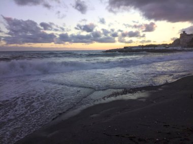 Gün batımında rüzgarlı bir günde deniz kıyısında kayalar ve binalarla kıyıya vuran dalgalar ve kumlar.