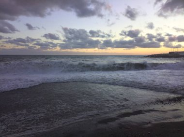 Gün batımında rüzgarlı bir günde deniz kıyısında kayalar ve binalarla kıyıya vuran dalgalar ve kumlar.