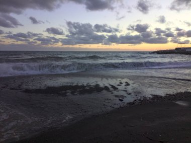 Gün batımında rüzgarlı bir günde deniz kıyısında kayalar ve binalarla kıyıya vuran dalgalar ve kumlar.