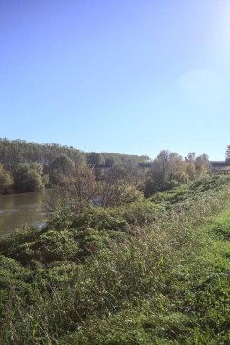 Nehir, kıyılarının kıyısındaki toprak setlerin arasında güneşli bir günde nehrin üzerinden geçen bir köprü ile İtalya 'nın kırsalında, toprak setin tepesindeki bir patikadan görülebiliyor.