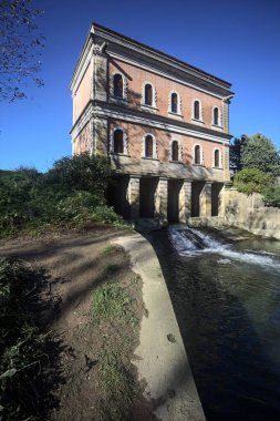 İtalya 'nın kırsal kesimindeki nehir kıyısındaki bir ağaç plantasyonundan görülen bir barajın üzerindeki tarihi bina.