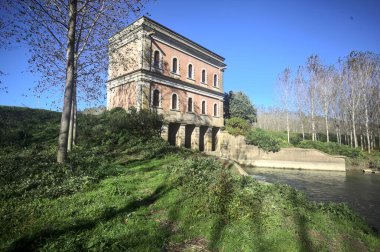 İtalya 'nın kırsal kesimindeki nehir kıyısındaki bir ağaç plantasyonundan görülen bir barajın üzerindeki tarihi bina.
