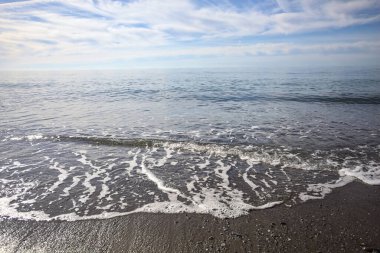 Kuma vuran dalgalarla kıyı şeridi ve güneşli bir günde ufukta durgun bir deniz.
