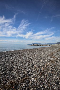 Güneşli bir günde kumsalda çakıl taşları ve arka planda kayalıklar vardı.