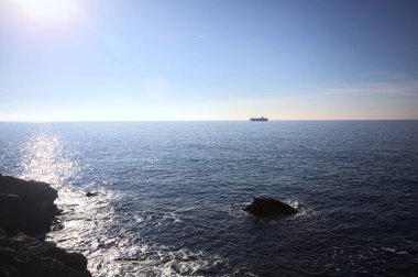Güneşli bir günde deniz, kayalara vuran dalgalar ve güneşli bir günde tepeden görünen bir kargo gemisi.