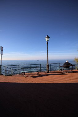 Yaşlı adam deniz kenarında bir bankta oturuyor gündüz vakti bir sokak lambasının yanında.