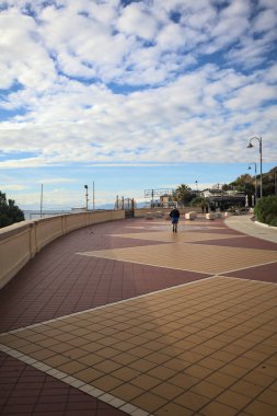 Cenova, İtalya - 2ND Aralık 2025 - Palmiyeler ve çiçek yataklarıyla çevrili deniz kenarı Corso Italia kaldırımı, insanların yürüdüğü sahil beldesi ve gök mavisi korkulukların arkasında, kışın bulutlu bir günde