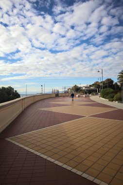 Cenova, İtalya - 2ND Aralık 2025 - Palmiyeler ve çiçek yataklarıyla çevrili deniz kenarı Corso Italia kaldırımı, insanların yürüdüğü sahil beldesi ve gök mavisi korkulukların arkasında, kışın bulutlu bir günde