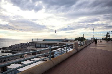 Cenova, İtalya - 2ND Aralık 2025 - Palmiyeler ve çiçek yataklarıyla çevrili deniz kenarı Corso Italia kaldırımı, insanların yürüdüğü sahil beldesi ve gök mavisi korkulukların arkasında, kışın bulutlu bir günde